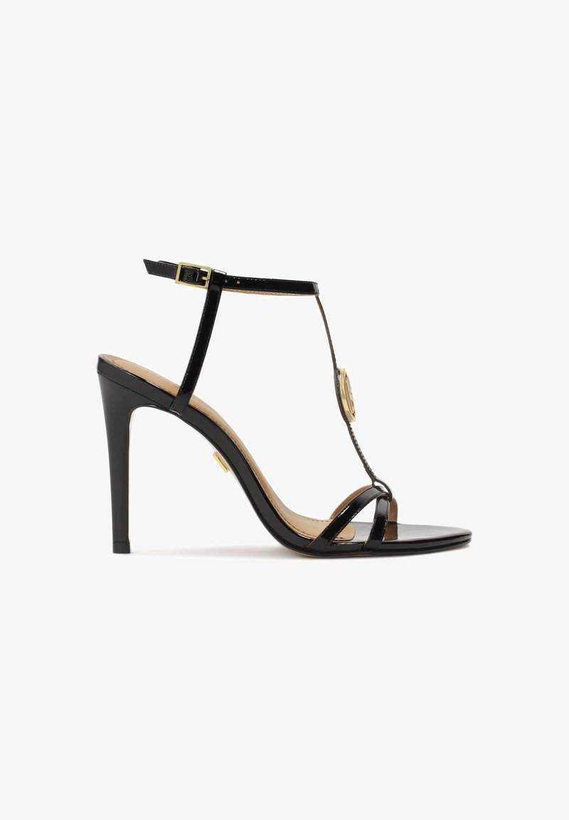 Černé lakované kožené sandály na vysokém podpatku s designem T-strap, nastavitelným zapínáním na kotníku a zlatým akcentem uprostřed.