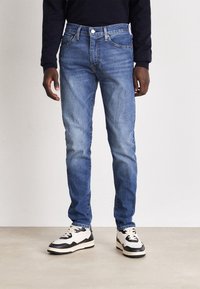 Blå denimjeans med slim fit, med midja i normalhöjd, fem fickor och diskret blekning. Kombinerat med svart och vita sneakers.