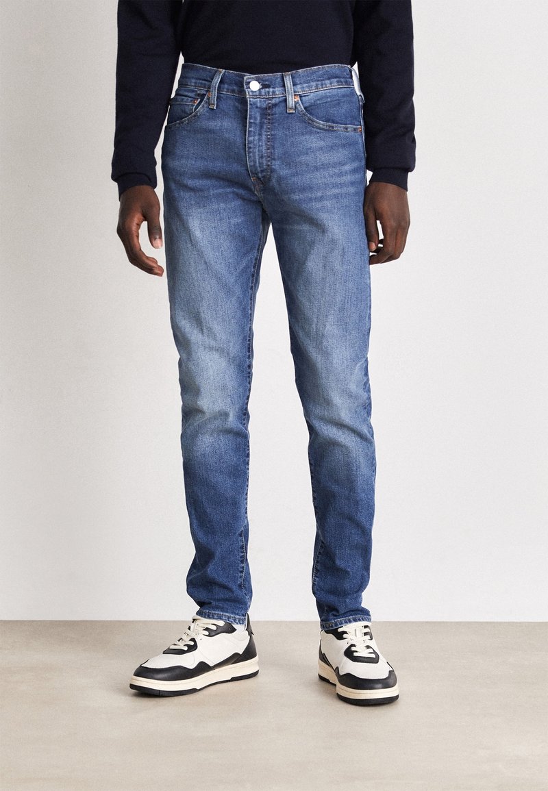 Blå denimjeans med slim fit, med midja i normalhöjd, fem fickor och diskret blekning. Kombinerat med svart och vita sneakers.