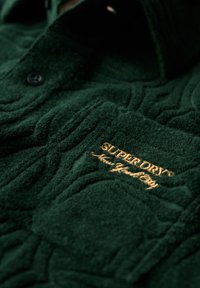 Donkergroen fleeceshirt met een textuurpatroon, voorzien van een borstzak en een geborduurd logo in goud. Zwarte knoopsluitingen zichtbaar.