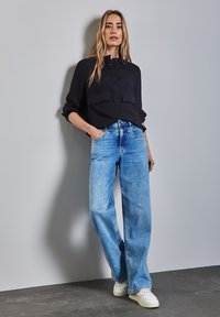 Camicia nera con bottoni e due tasche frontali, abbinata a jeans a gamba larga blu chiaro e sneakers bianche su un pavimento grigio testurizzato.