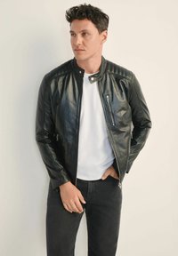 Czarna skórzana kurtka z stójką, pikowanymi detalami na ramionach, z przodu na zamek błyskawiczny i bocznymi kieszeniami. Nosi się nad białą koszulką.