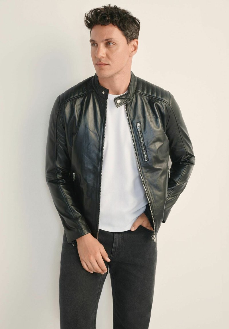 Czarna skórzana kurtka z stójką, pikowanymi detalami na ramionach, z przodu na zamek błyskawiczny i bocznymi kieszeniami. Nosi się nad białą koszulką.