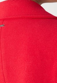 Tissu de manteau texturé rouge vif avec une petite étiquette rectangulaire noire et argentée près du col sur le haut du dos.