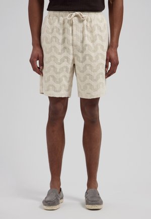 Man die beige gebreide korte broek met golfpatroon, trekkoord in de taille, donker shirt en grijze instapschoenen draagt, staand tegen een effen achtergrond.