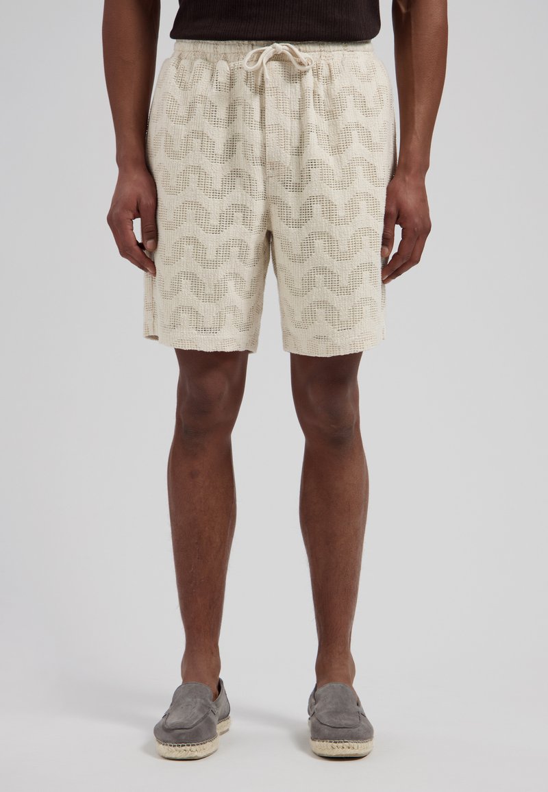 Homme portant un short en tricot beige avec motif ondulé, taille à cordon, haut sombre et chaussures à enfiler grises, debout devant un fond uni.