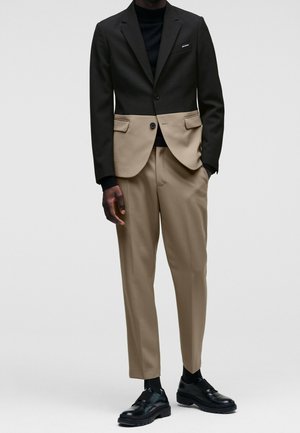 Homme portant un blazer bicolore noir et beige avec un pantalon beige et des chaussures habillées noires, debout devant un fond uni.