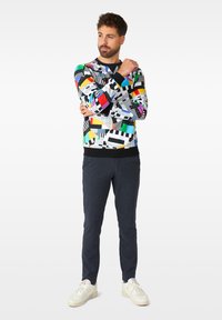 Wielokolorowy sweter w wzory geometryczne, zestawiony z fakturowanymi ciemnymi spodniami i białymi sneakersami. Kluczowe cechy to odważne kolory i kontrast.