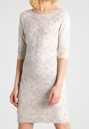 Femme portant une robe beige clair longueur genoux avec des manches trois-quarts et des motifs floraux en dentelle en relief à l'avant.