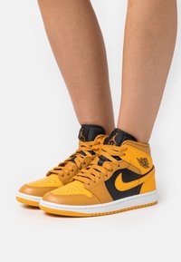 Osoba oblečená v žlutých a černých vysokých botách Nike Air Jordan s oranžovými tkaničkami a bílými podrážkami, stojící na bílém pozadí.