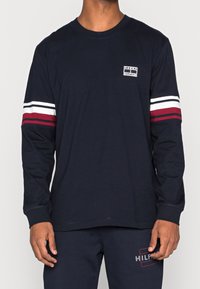 Marineblaues Langarmshirt aus Baumwolle mit weißen und bordeauxfarbenen Streifen an den Ärmeln, kleinem Logo-Patch auf der Brust und rundem Ausschnitt.