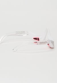 Białe sportowe okulary przeciwsłoneczne z przezroczystymi ramionkami i jasnoróżowymi akcentami. Zawierają oznaczenia marki oraz znaki bezpieczeństwa na wewnętrznych ramionach.