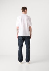 White short-sleeved shirt, relaxed fit, worn with dark blue denim jeans, standing on a plain background. Visible tattoo on arm.