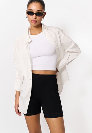 Kobieta w czarnych krótkich spodenkach kolarskich, białym krótko skrojonym topie na ramiączkach, dużej białej koszuli z guzikami i czarnych wąskich okularach przeciwsłonecznych na białym tle.