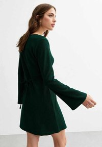 Abito verde a coste con maniche lunghe svasate e cintura con lacci. Presenta un orlo dritto, creando una silhouette aderente sul retro.