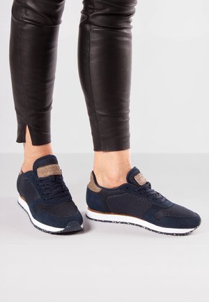 Person iført sorte læderbukser og marineblå mesh-sneakers med beige accenter, stående på en grå gulv med hvid baggrund.