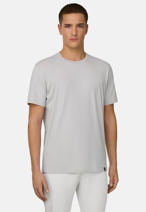 Giovane in piedi, indossa una maglietta a maniche corte grigio chiaro unita e pantaloni bianchi su uno sfondo semplice.