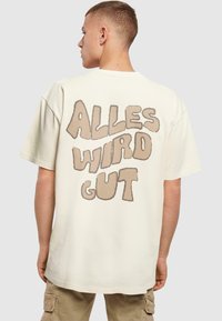 Cremefarvet bomulds t-shirt med et stort, lysebrunt boble-bogstav grafisk motiv på ryggen, der siger "ALLES WIRD GUT." Løs pasform, korte ærmer.