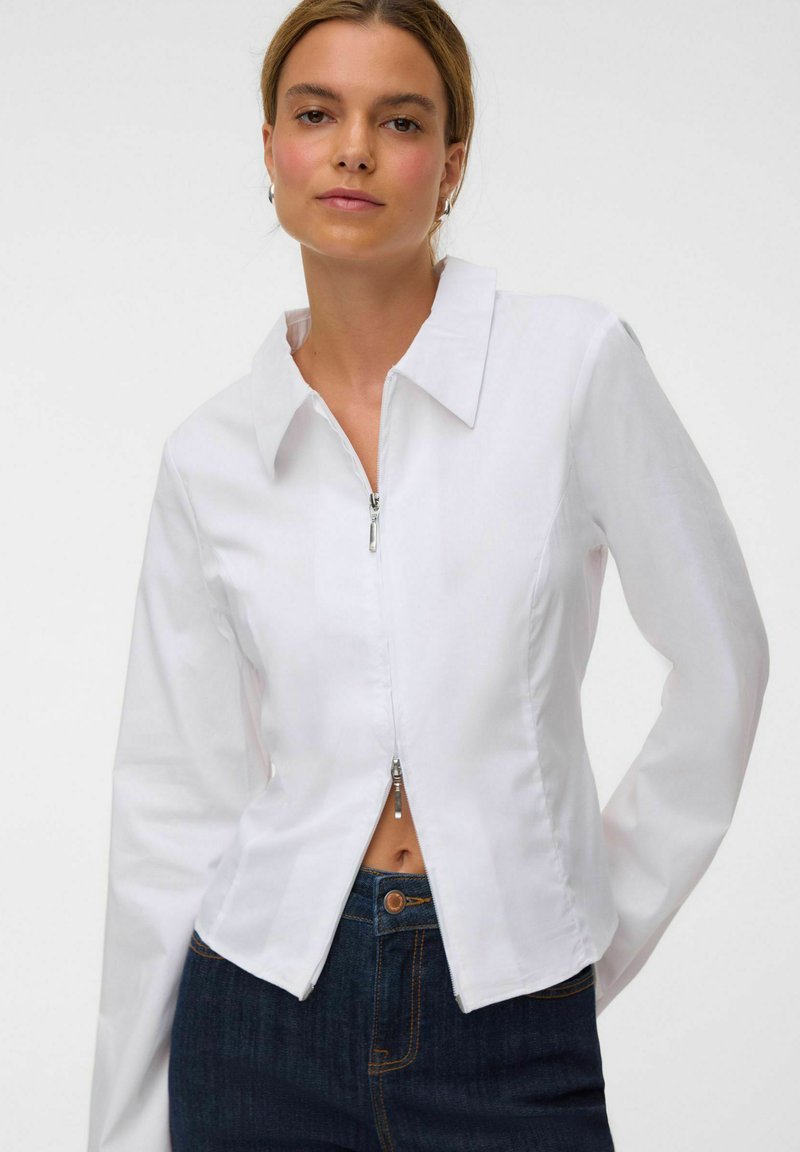 Femme portant une chemise blanche ajustée à manches longues avec une double fermeture éclair, associée à un jean taille haute bleu foncé sur un fond uni.
