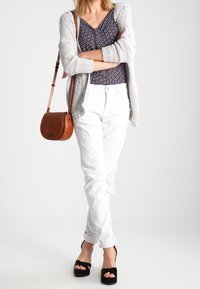 Jasnoszara, dzianinowa kamizelka na wzorzystej granatowej bluzce, białe spodnie o teksturowych detalach, czarne buty na platformie oraz brązowa torebka na ramię.