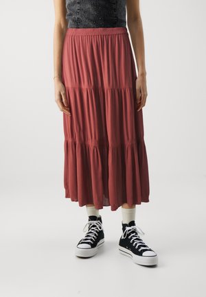 Vrouw die een lange, gelaagde roestkleurige rok, witte sokken en zwarte hoge sneakers draagt, staand tegen een effen achtergrond.