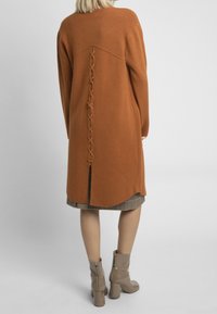 Długi pomarańczowy sweter z fakturowanym tyłem z ozdobnym sznurowaniem. Ma wysokie rozcięcie, noszony na kratkowaną spódnicę i zestawiony z beżowymi botkami na obcasie.