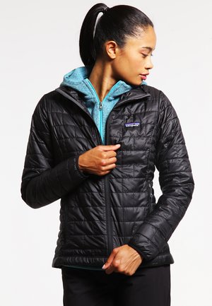 Giacca trapuntata nera con zip frontale, dotata di cappuccio foderato in pile blu teal. Il tessuto appare liscio, con una texture leggera.