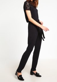 Černý overal s krajkovými krátkými rukávy, úzkým pasem svázaným páskem a rovnými nohavicemi. Nosený s černými lesklými mokasíny.