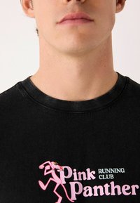 Must t-särk mustast puuvillast, millel on roosa Panteriga graafika ja tekst "Pink Panther RUNNING CLUB". Ümar kaelus ja pehme tekstuur.