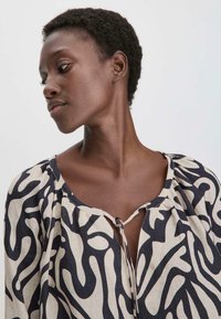 Maglietta a maniche lunghe in lino beige con motivo astratto nero. Caratterizzata da un collo rotondo e un dettaglio con fiocco al collo, mostra una vestibilità rilassata.