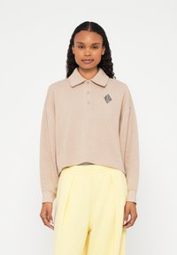 Polo bege de manga comprida com colarinho, três botões e um logótipo geométrico preto no lado esquerdo do peito, combinado com calças largas amarelo-claro.