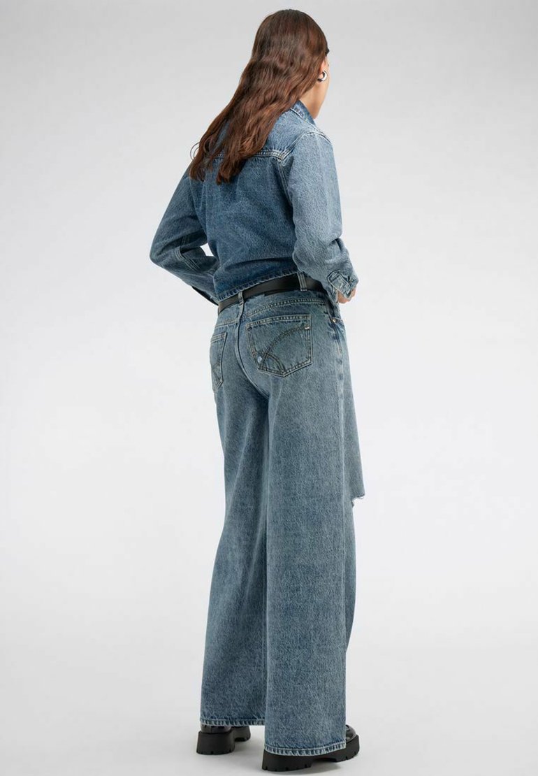 Giacca di jeans blu con una texture leggermente sbiadita abbinata a jeans blu a vita alta. Presenta una cintura nera e scarpe nere chunky.