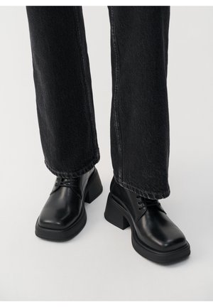 Chaussures noires en cuir à lacets avec un talon épais et chunky et un bout arrondi, portées avec un pantalon en denim noir ample.