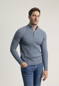 Suéter de punto trenzado de color azul claro con un cuello alto y tres botones. Puños y dobladillo acanalados. Lleva puesto unos jeans de color azul oscuro.