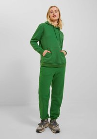 Green hooded sweatshirt with front pocket and matching sweatpants. Soft fabric with a smooth texture. Worn with gray sneakers.