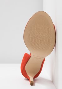 Chaussure à talon haut en daim rouge placée semelle contre un mur blanc, montrant la semelle texturée et le talon fin. Taille 37, fabricant San Marina.