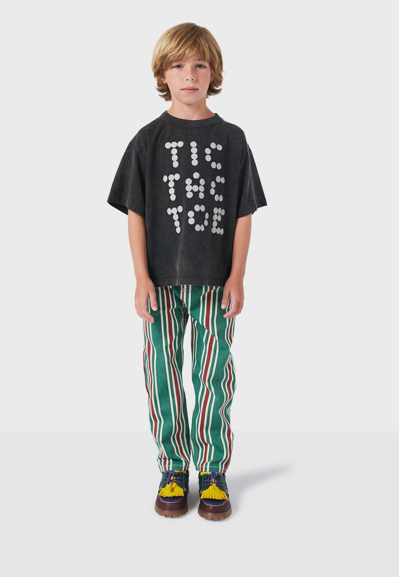 Zwart T-shirt met een wit stippenpatroon dat "TIC TAC TOE" spelt, gecombineerd met groene broek met rode en witte verticale strepen, en meerkleurige schoenen.