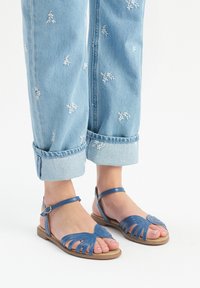 Niebieskie skórzane sandały mają wycięcia w zdobieniach w kształcie liści, regulowany pasek na kostce oraz płaską podeszwę, w połączeniu z podwiniętymi jasnoniebieskimi dżinsami.