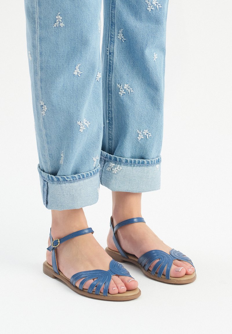 Niebieskie skórzane sandały mają wycięcia w zdobieniach w kształcie liści, regulowany pasek na kostce oraz płaską podeszwę, w połączeniu z podwiniętymi jasnoniebieskimi dżinsami.