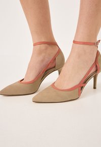 Scarpe beige in suede con tacco alto e punta affilata, con un accento corallo attorno all'apertura e una cinghia alla caviglia regolabile per una vestibilità sicura.