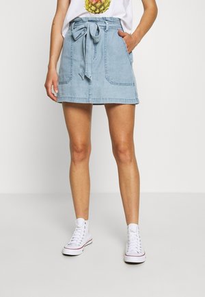 Vrouw die een lichtblauwe denimrok met zakken aan de voorkant en een strikceintuur draagt, witte hoge sneakers en een wit T-shirt met een ananasprint.