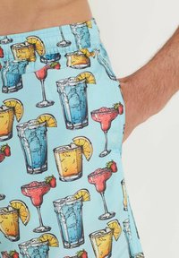 Shorts de bain avec un fond bleu clair présentant un motif de cocktails colorés, comprenant des verres avec des boissons, des tranches de fruits et des accents.