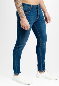 Vaqueros ajustados de color azul con textura descolorida, cinco bolsillos y costuras en contraste, combinados con zapatillas blancas sobre un fondo liso.