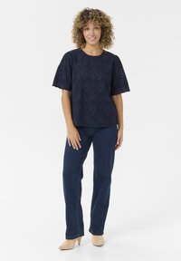 Haut bleu marine brodé avec des manches courtes à volants, présentant des motifs découpés, associé à un jean foncé et des chaussures à talons beiges.