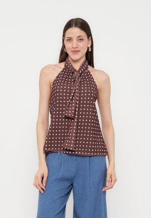 Donna che indossa un top marrone senza maniche con pois bianchi e un nodo sul davanti, abbinato a pantaloni blu ampi, in piedi su uno sfondo chiaro.