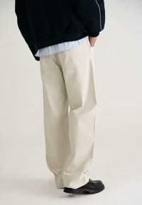Pantaloni bej cu picioare largi din bumbac, prevăzuți cu buzunare la spate, asortați cu o cămașă albastră cu dungi și încălțăminte chunky negre. Design simplu și funcțional.