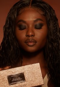 Rektangulært makeup-palette i rosagulv som holdes i hånden, med en glatt overflate og gravert etikett, mot en varm, nøytral bakgrunn.