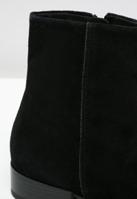 Bottine en daim noir avec couture visible et petit talon empilé sur fond blanc.