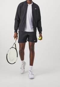 Černá sportovní bomber bunda, bílá termoaktivní košile, černé šortky s logem, bílé tenisové boty, držící tenisovou raketu a zelený míček.