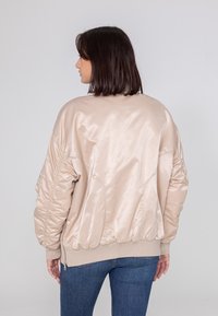 Beżowa satynowa kurtka bomberka z prążkowanym dołem i mankietami, z marszczonymi szwami na ramionach i zamykaną na zamek kieszenią na rękawie.